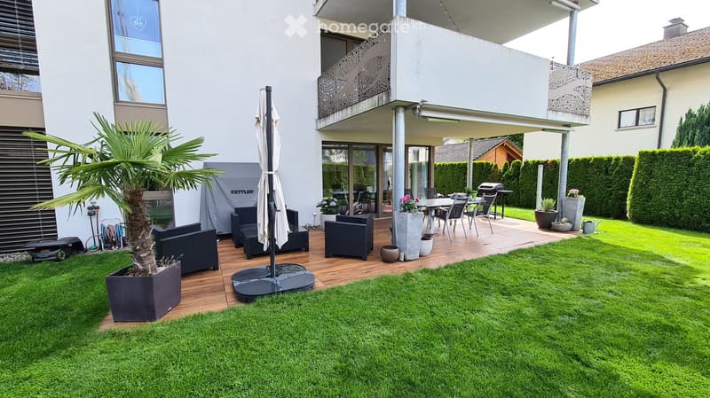 Well-maintained garden with a wooden deck, outdoor seating area, umbrella, sofa, ottoman, and flower pots.