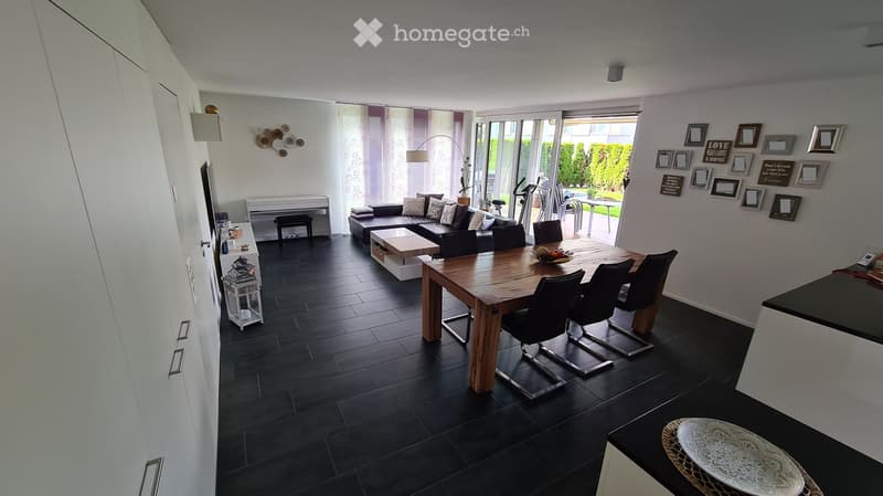 Modern living and dining area with dark tiled flooring, a black sofa, a wooden coffee table, a dining table with chairs, and a small round stool.