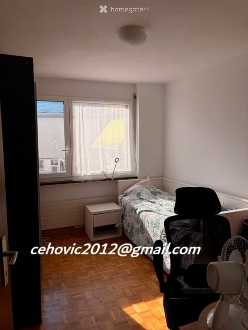 Cozy bedroom with wooden flooring, a single bed with patterned bedding, a nightstand, a chair, and a fan near the window.