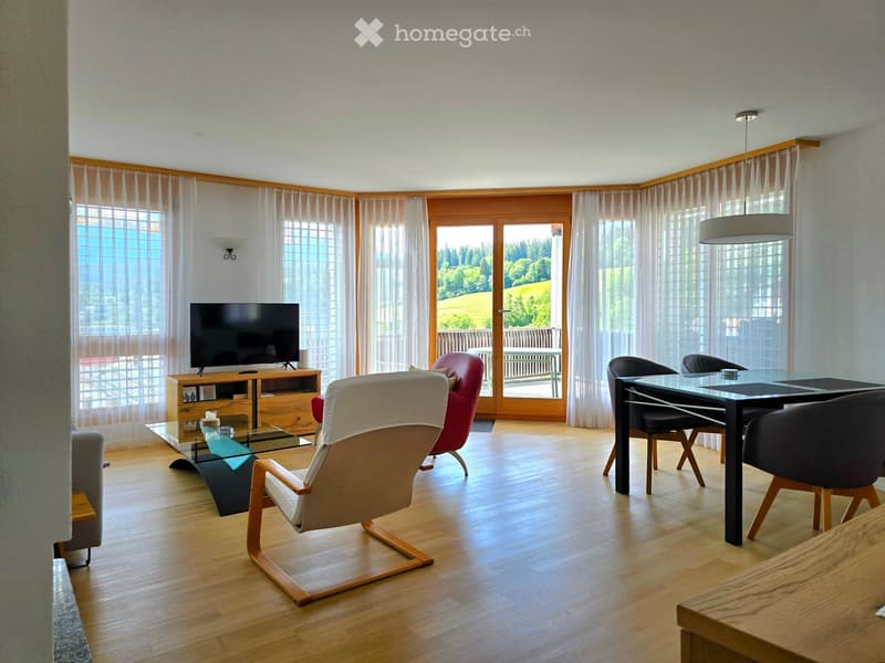 Modern living room with wooden flooring, large windows, and a view of greenery. Features include a sofa, chairs, a glass coffee table, a TV on a wooden stand, and a dining table with chairs.
