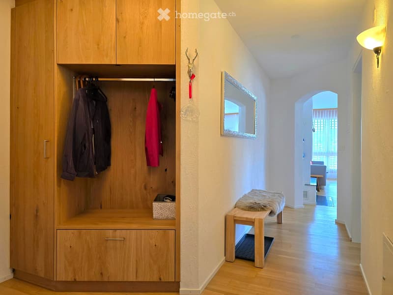 A modern hallway with wooden flooring, a built-in coat closet, a small bench with a fur throw, and a mirror on the wall.