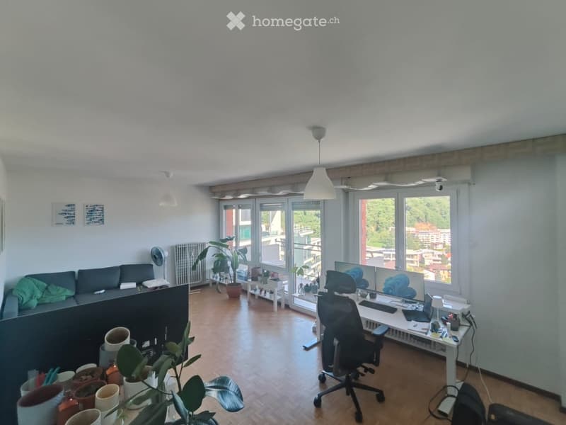 A modern, well-lit room with a sofa, office chair, desk, laptop, and potted plants. Large windows offer a view of the city.