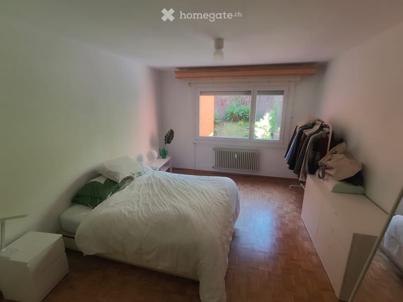 Cozy bedroom with wooden flooring, a double bed with white bedding, a nightstand with a lamp, a mirror, and a radiator under the window. Clothes are hanging on a rack near the window.