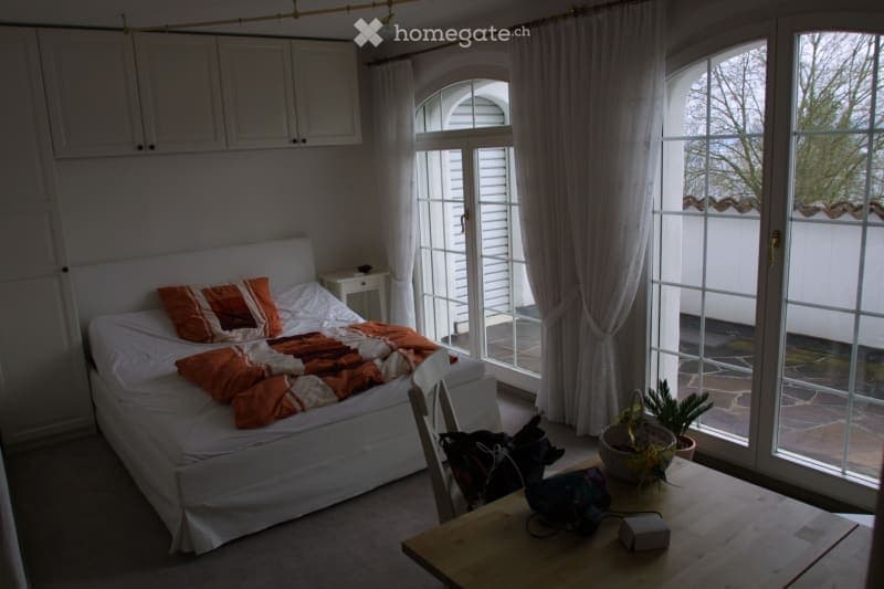 Bright bedroom with a white bed, orange pillows and blanket, a nightstand, a chair, a small table with a plant, and large windows leading to a balcony.