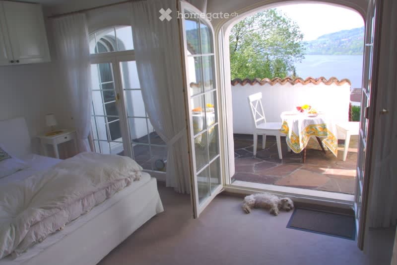 Bright bedroom with large windows and an open door leading to a balcony overlooking a scenic lake view. The room features a neatly made bed, a nightstand with a lamp, and a small dog resting on the floor.