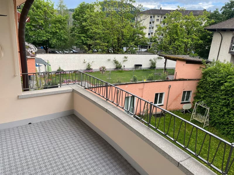 Balcony with metal railing, tiled floor, overlooking a green garden area with trees, a small table, and a swing chair.