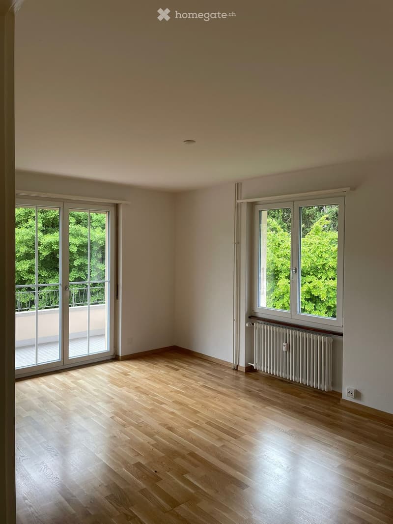 Empty room with light wooden flooring, white walls, large windows, and a radiator. No furniture or equipment present.