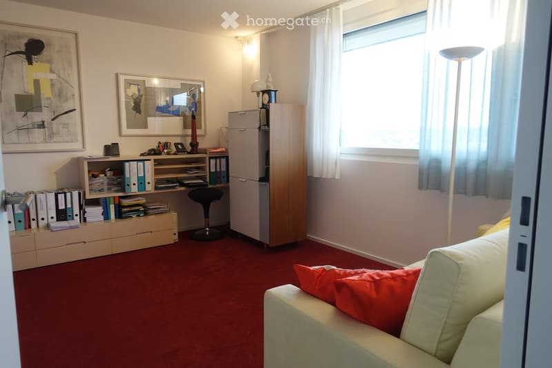 A well-lit room with a red carpet, a white sofa with red cushions, a wooden desk with binders and books, a black stool, a filing cabinet, and a mirror on the wall.