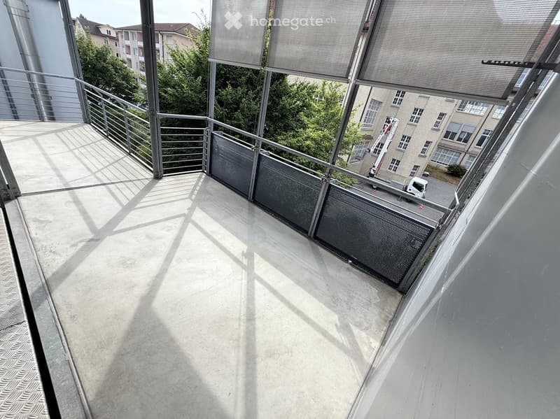 Modern balcony with metal railings, concrete floor, and large windows overlooking a residential area with trees and buildings.