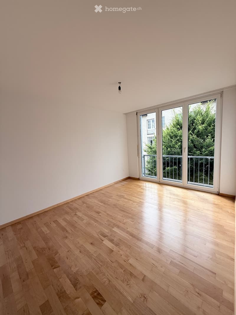 Empty room with light wooden flooring and white walls, featuring large sliding glass doors leading to a balcony with a view of greenery.