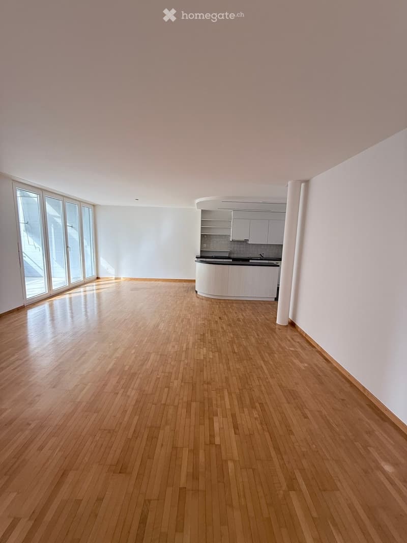 Empty room with light wooden flooring, white walls, and a small kitchen area with cabinets and a countertop visible in the background. Large windows allow natural light to enter.