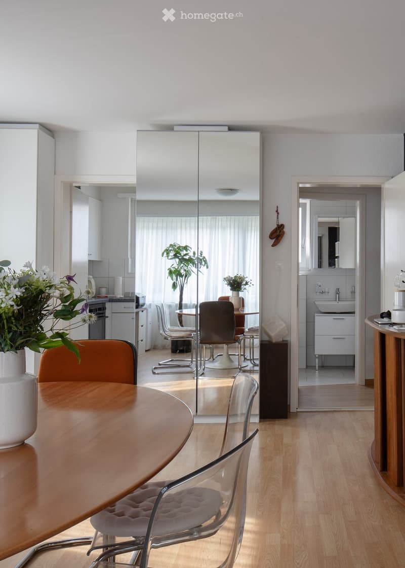 A modern, well-lit dining area with a wooden table, chairs, a large mirror, and a vase with flowers. The room features light wooden flooring and white walls, with a view into a kitchen area.