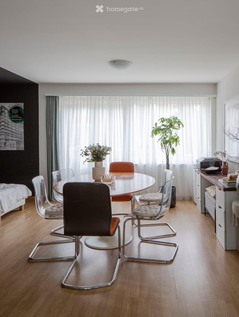 Modern, well-lit room with a round dining table surrounded by chairs, a large plant, a bed, and a cabinet with a printer on top.