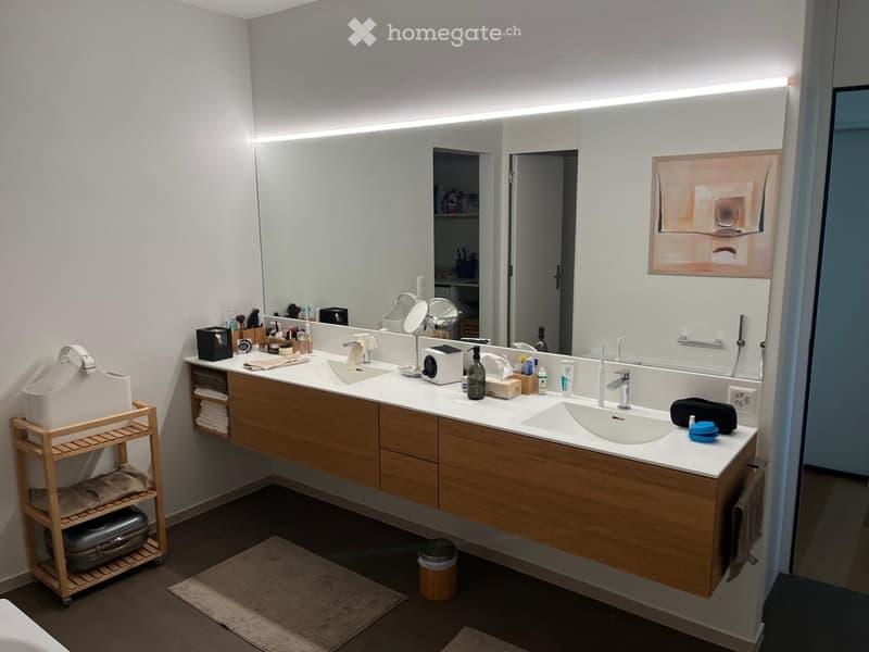Modern bathroom with double sink vanity, large mirror with built-in lighting, various toiletries, and a small wooden shelf holding a handbag and luggage.