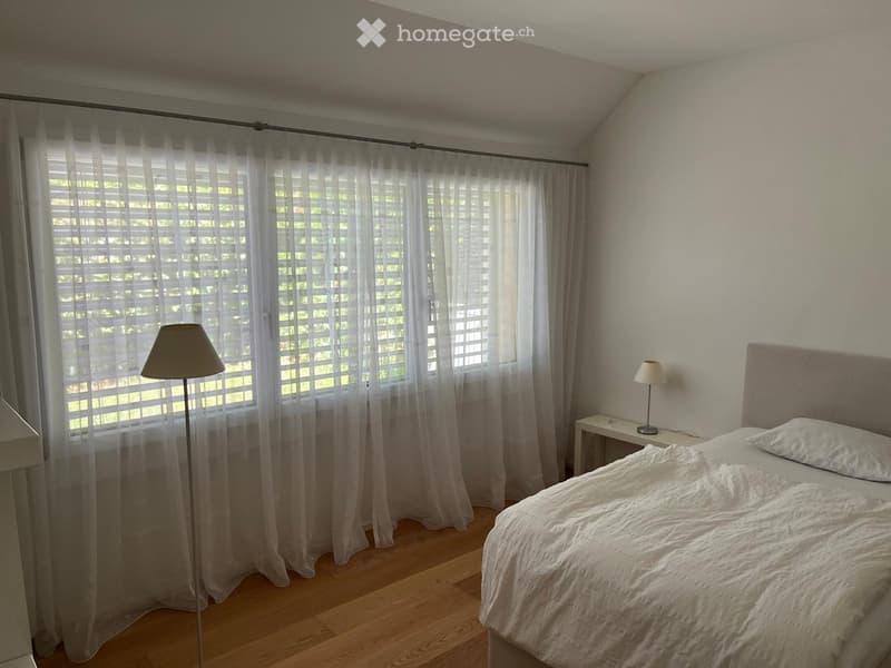 Bedroom with a neatly made bed, nightstand, and lamps. Large window with sheer curtains allows natural light to fill the space.
