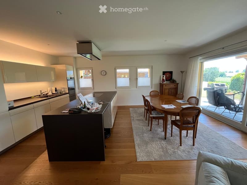 Open-plan kitchen and dining area with light-colored cabinets, a central island, a dining table with chairs, and a sofa visible in the foreground.