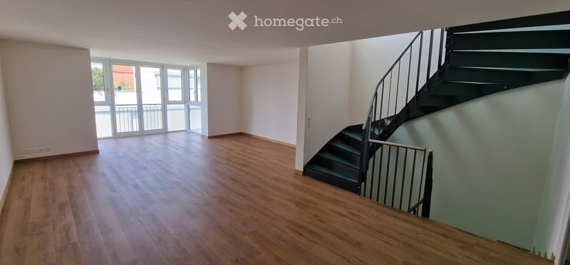 Empty room with wooden flooring, white walls, and a staircase leading upwards. Large windows with blinds provide natural light.