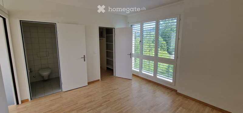 Empty room with wooden flooring, large windows with blinds, and an open door leading to a small bathroom with a toilet.