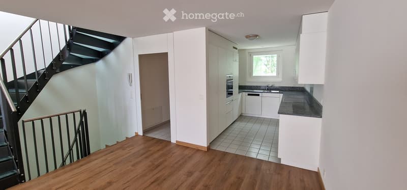 Modern, well-lit interior with a wooden floor, staircase leading upwards, and a compact kitchen area featuring white cabinets, a sink, and an oven.