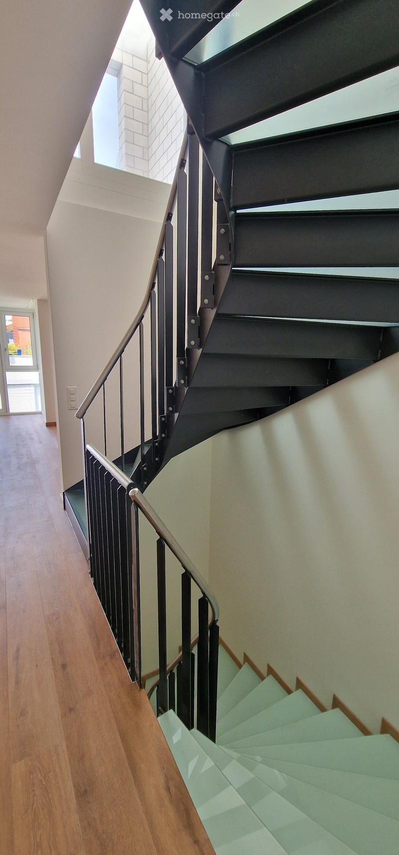 Modern staircase with wooden steps, metal railing, and glass panels, leading to a bright hallway with wooden flooring and white walls.