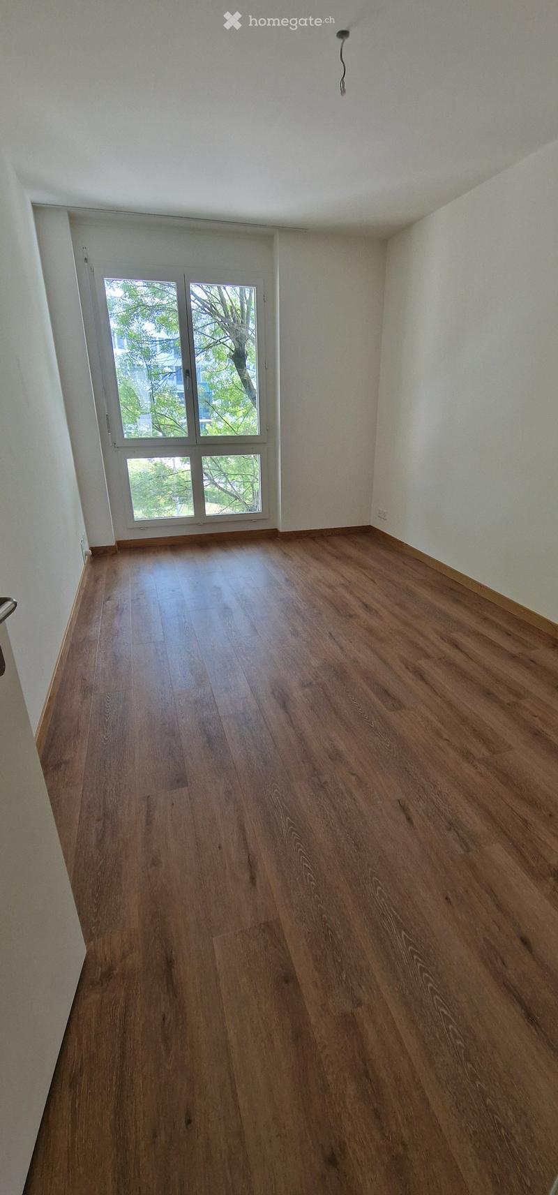 Empty room with wooden flooring, white walls, and large windows with a view of trees outside.