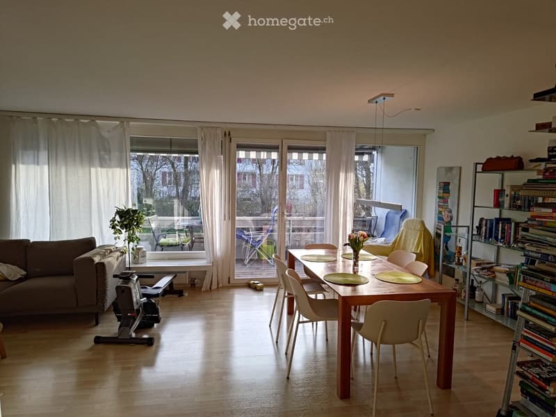 Bright living and dining area with wooden flooring, large windows, a sofa, dining table with chairs, bookshelf, and exercise bike.
