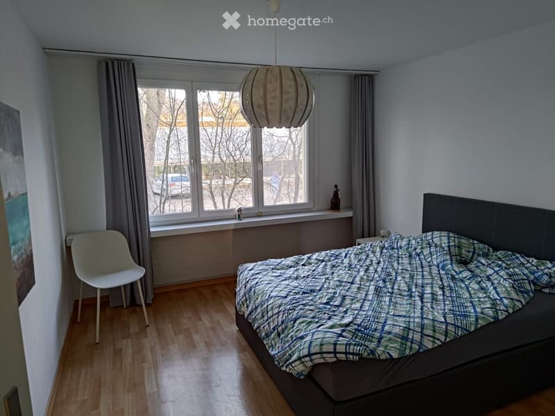 Bedroom with wooden floor, large window with curtains, bed with headboard and plaid blanket, white chair, and a hanging lamp.
