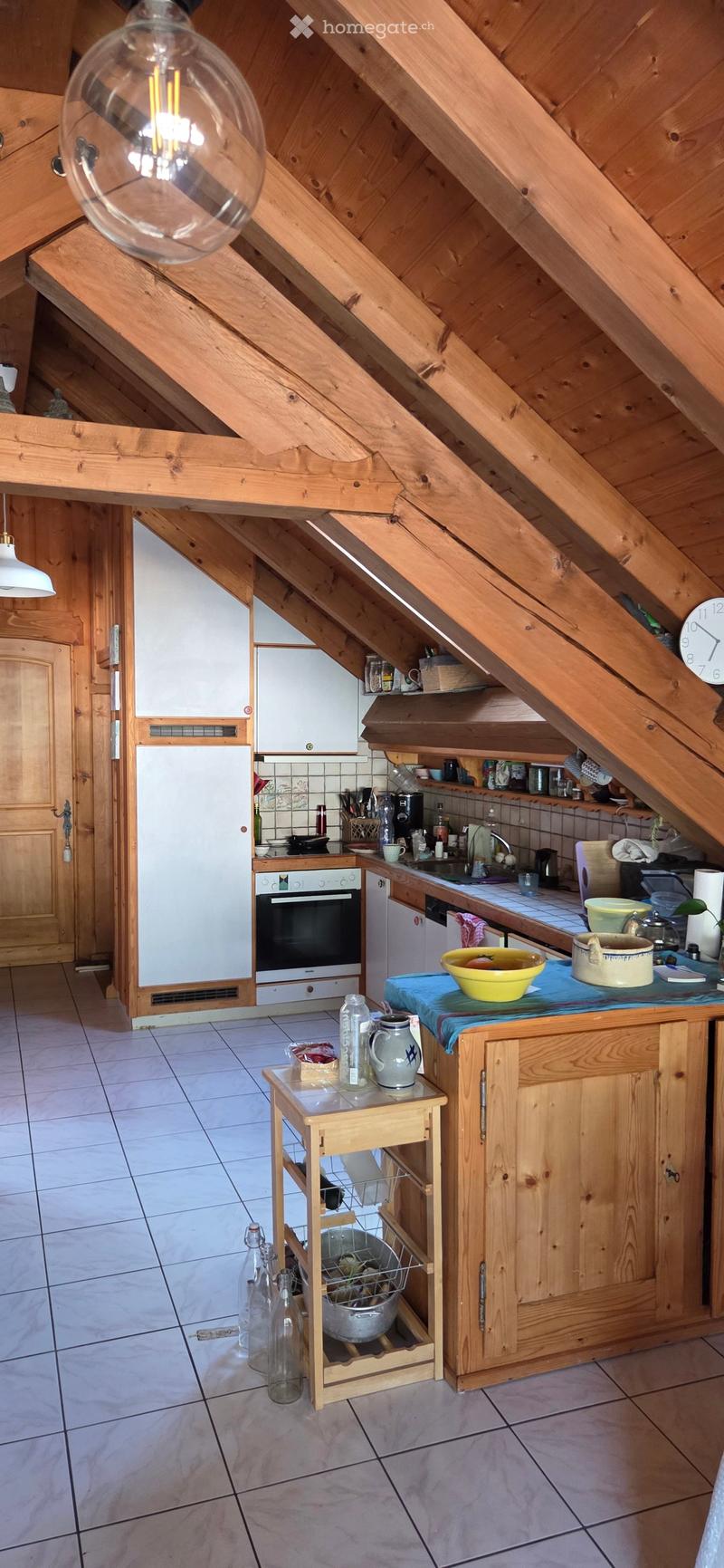 Cozy attic kitchen with wooden beams, white cabinets, and various appliances including a refrigerator, oven, and dishwasher. The space features a small wooden table with bottles and a bowl on the counter.