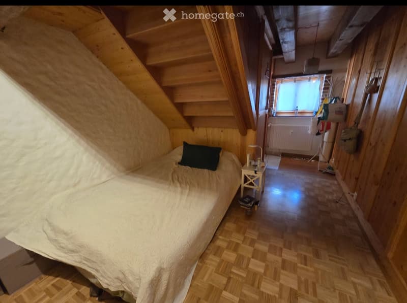Cozy attic bedroom with wooden walls and slanted ceilings, featuring a single bed, nightstand, and a view of a hallway leading to another room.
