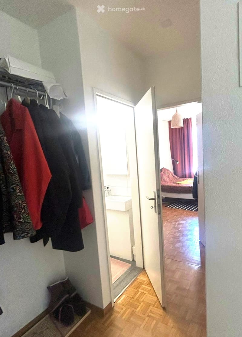 Hallway with wooden flooring, coat rack with hanging clothes, open door leading to a bedroom with a bed, red curtains, and a small sink area visible in the background.