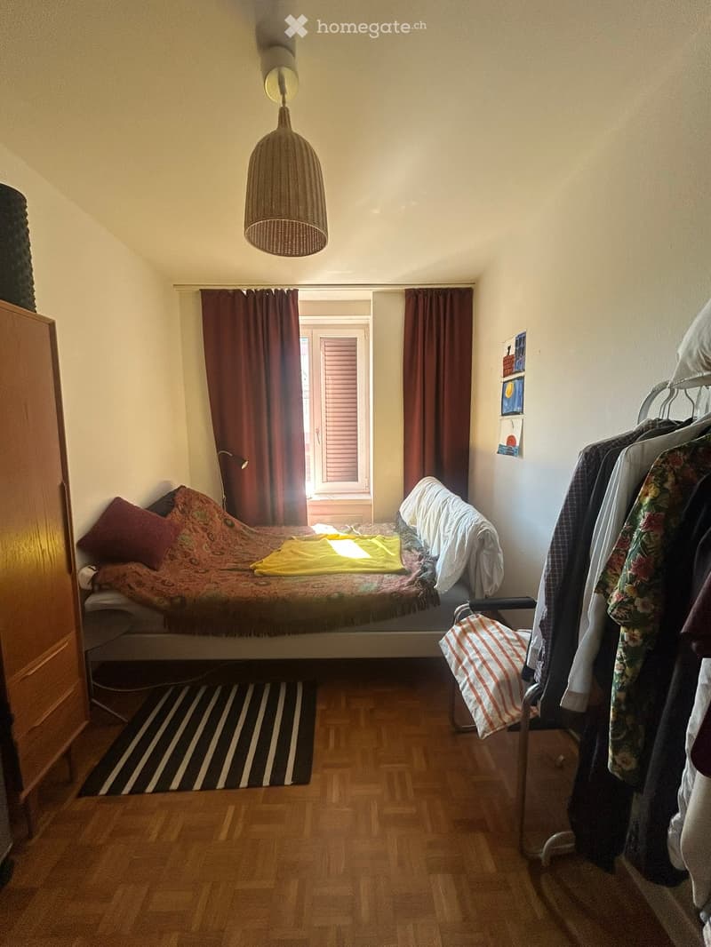 Cozy bedroom with wooden floor, bed with colorful bedding, wardrobe, and clothes hanging on a rack. Features a hanging lamp, striped rug, and artwork on the wall.