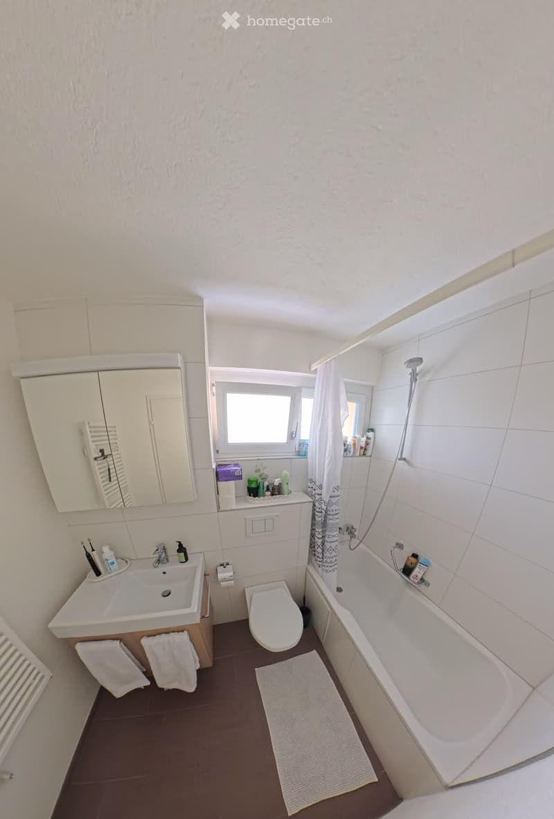 Modern bathroom with white tiles, a sink, toilet, bathtub, mirror, and a window. Includes towels, a shower curtain, and various toiletries on the sink and bathtub ledge.