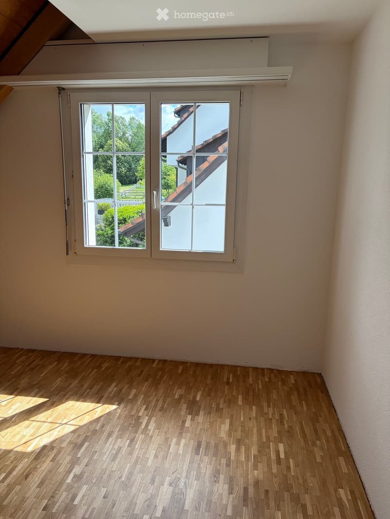 Empty room with wooden flooring and large windows showing a view of greenery and a neighboring building.