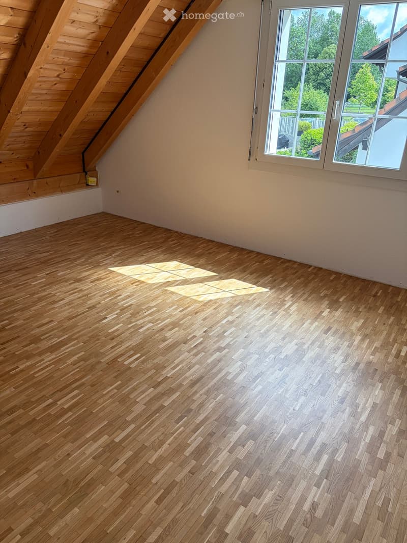 Empty attic room with wooden floor and slanted wooden ceiling, large window letting in natural light.