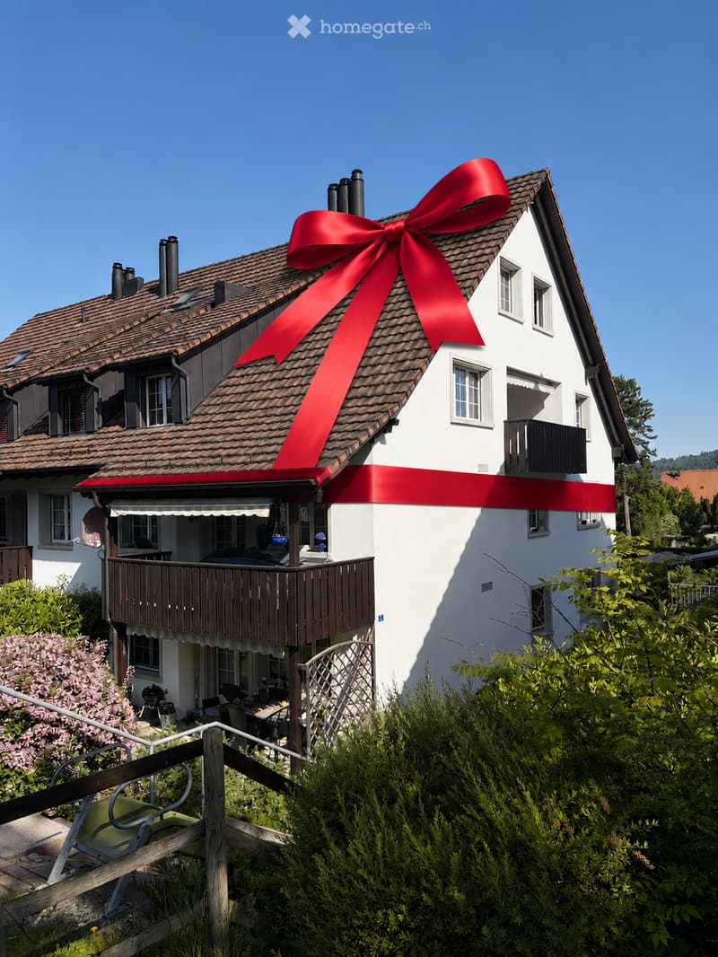 Two-story house with a red roof and white walls, featuring a large red ribbon on the roof, a balcony, and a well-maintained garden.