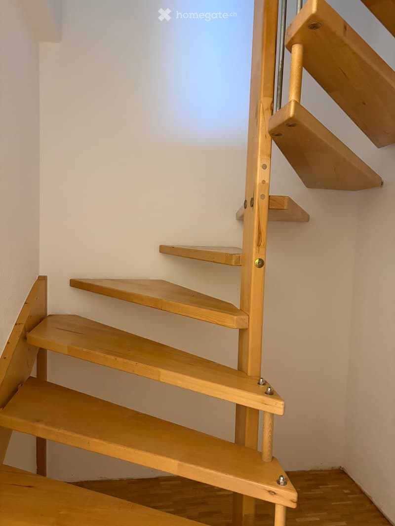 Wooden staircase with open shelves and a handrail, set against a white wall with a window above.