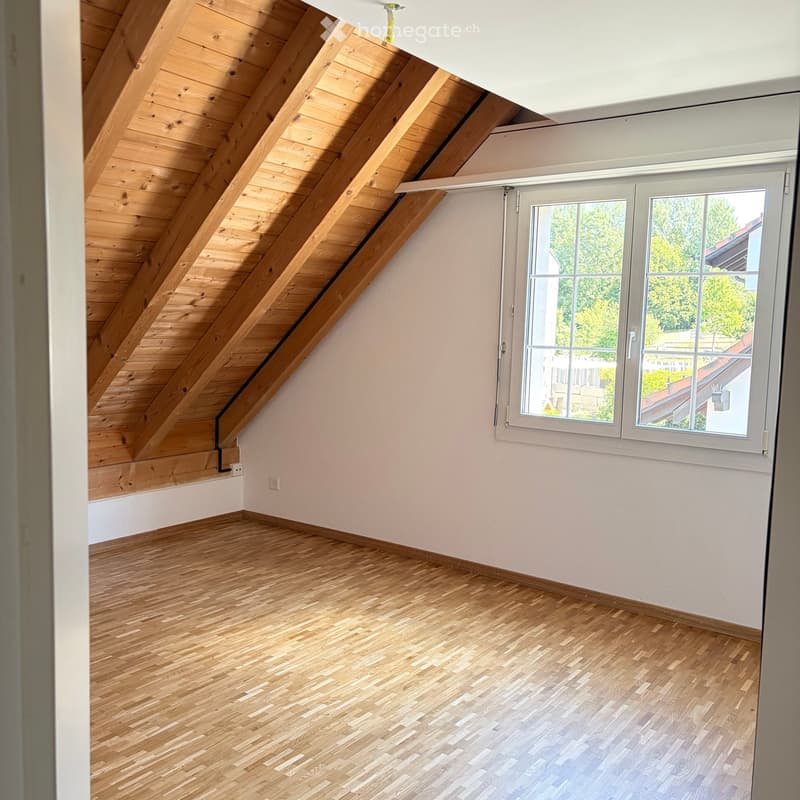 Empty attic room with wooden slanted ceiling, light wooden flooring, and a large window offering a view of greenery.