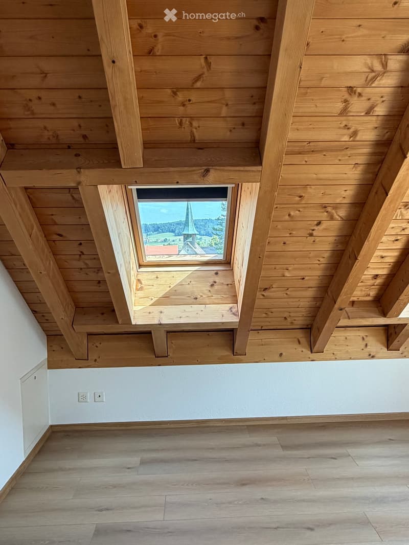 Empty room with wooden ceiling beams, exposed wooden floor, and a skylight window offering a view of a church and landscape.