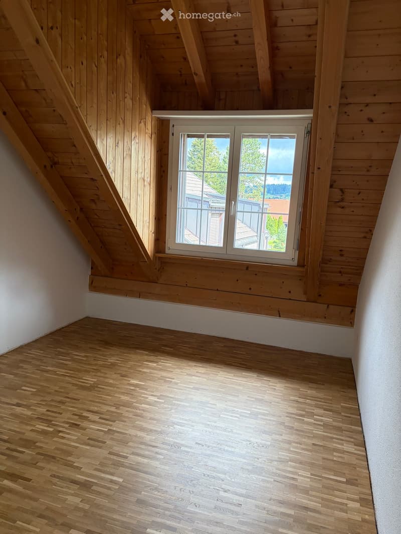 Empty attic room with wooden walls, ceiling beams, and a large window offering a view of the outside. The floor is made of light wooden planks.