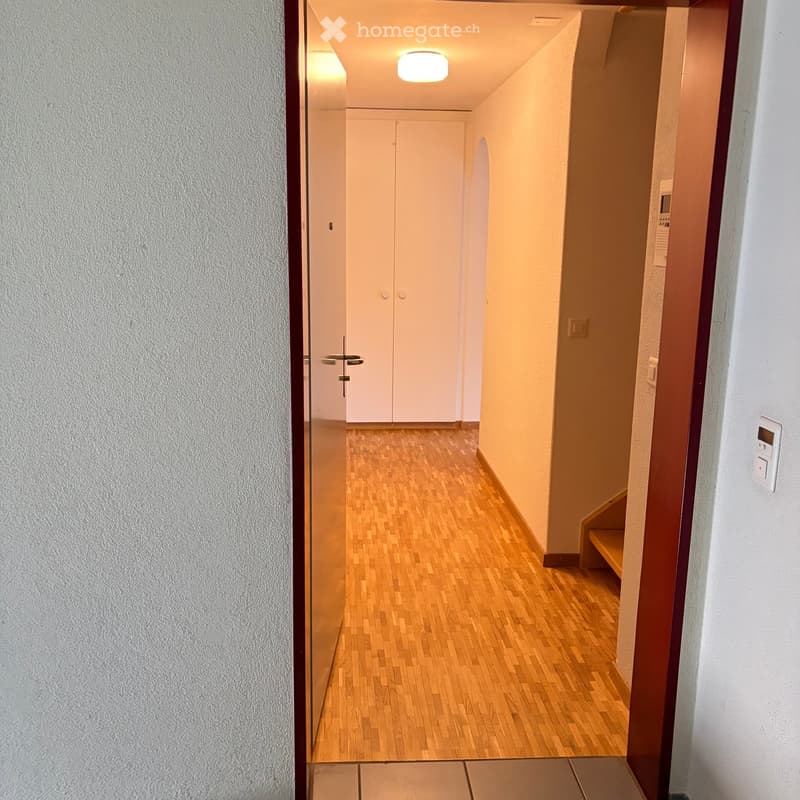 A narrow hallway with wooden flooring, white walls, and a closed double door at the end. A small wooden bench is visible on the right side.