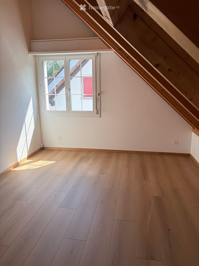 Empty attic room with wooden flooring, white walls, and a large window letting in natural light. No furniture or equipment is present.