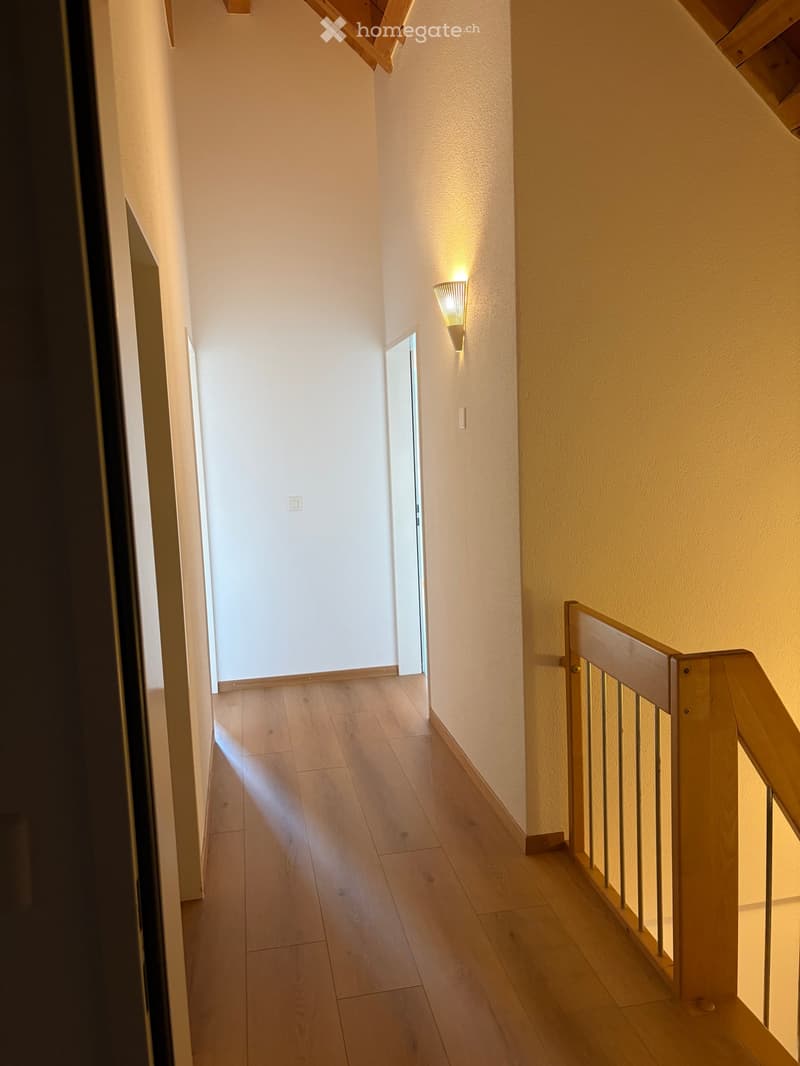 A narrow hallway with light wooden flooring, white walls, and a wooden staircase railing on the right side. A wall-mounted light fixture provides warm illumination, and a small open doorway leads to another room.
