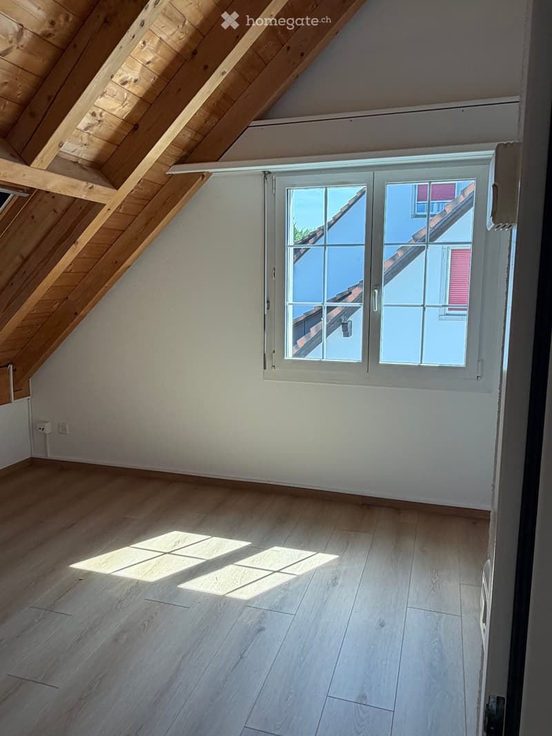 Empty attic room with wooden slanted ceiling, light wooden floor, and large window showing a view of the roof and sky.