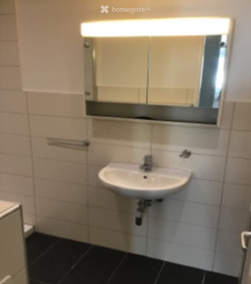 Modern bathroom with white tiled walls, black tiled floor, a white sink, a large mirror with built-in lighting, and a chrome faucet.