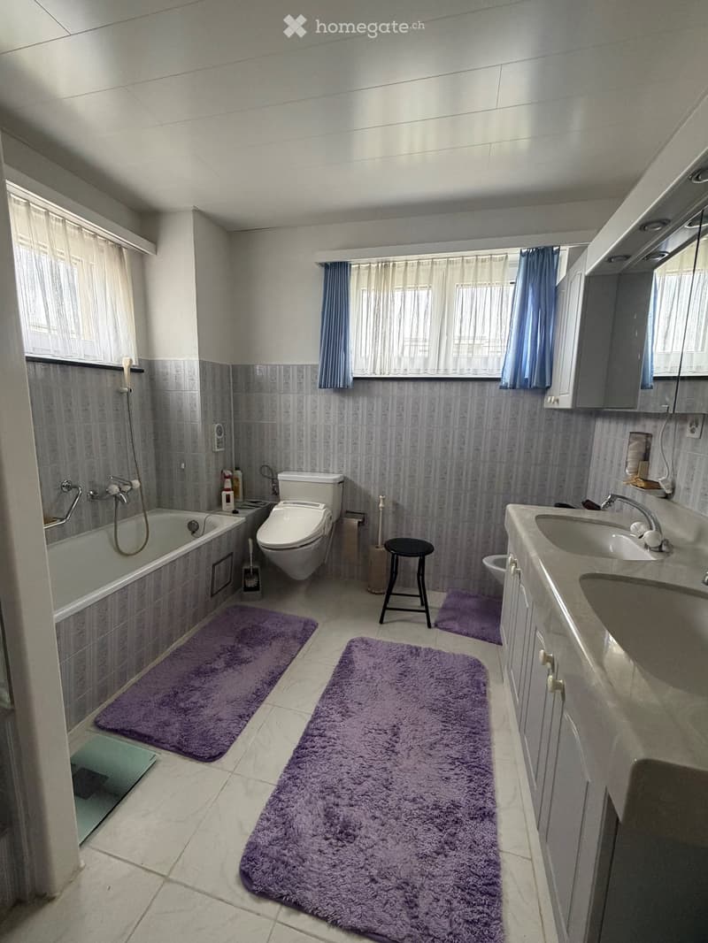 Modern bathroom with a bathtub, toilet, sink, and vanity cabinet. Features purple rugs, a small stool, and a window with curtains.