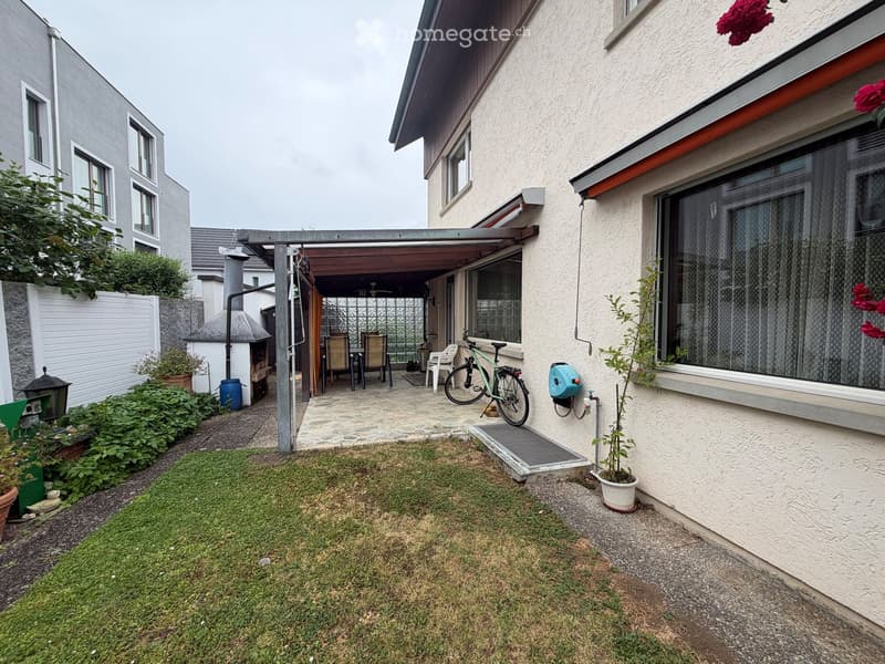 Garden area with a covered patio, outdoor furniture, bicycle, and small plants. The patio has a stone floor and is connected to the house via a small staircase.