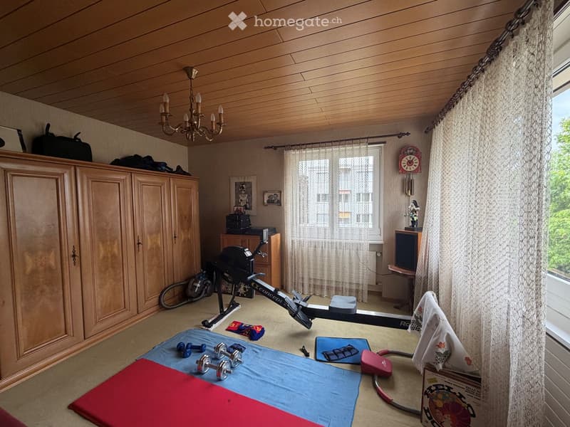 Well-lit room with wooden ceiling and walls, featuring exercise bike, dumbbells, cabinet, chandelier, and large window with curtains.