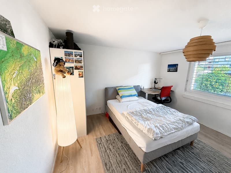 Cozy bedroom with a single bed, desk, chair, and a tall floor lamp. The room features a large window with a view of greenery, a map on the wall, and a small shelf with photos and a teddy bear.