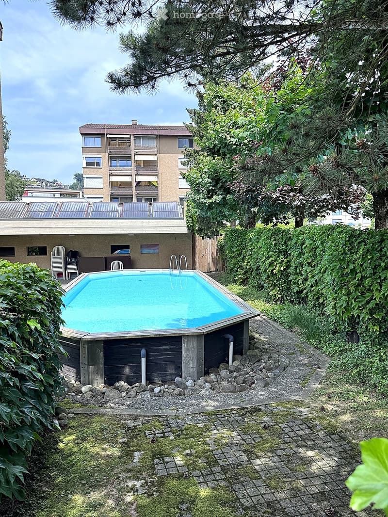 A small rectangular swimming pool surrounded by greenery, with a paved pathway and a multi-story building in the background.