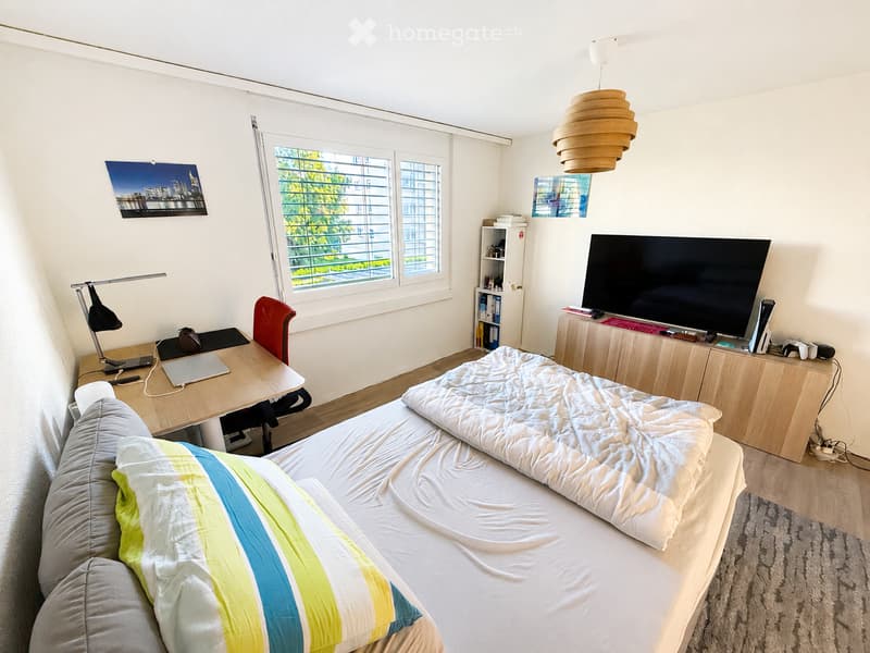 A well-lit bedroom with a bed, desk, chair, laptop, lamp, bookshelf, and a large screen. The room features large windows with blinds and a modern aesthetic.